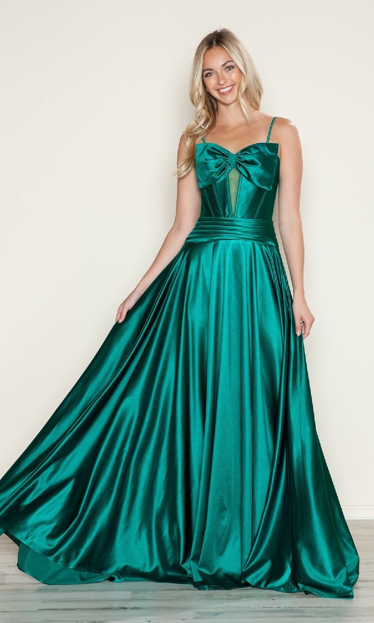 A woman wears the Bow-Front Long Corset Prom Dress (Poly USA 9702) in emerald, featuring shiny satin fabric, spaghetti straps, a fitted bodice, and a large bow detail, standing against a light background.